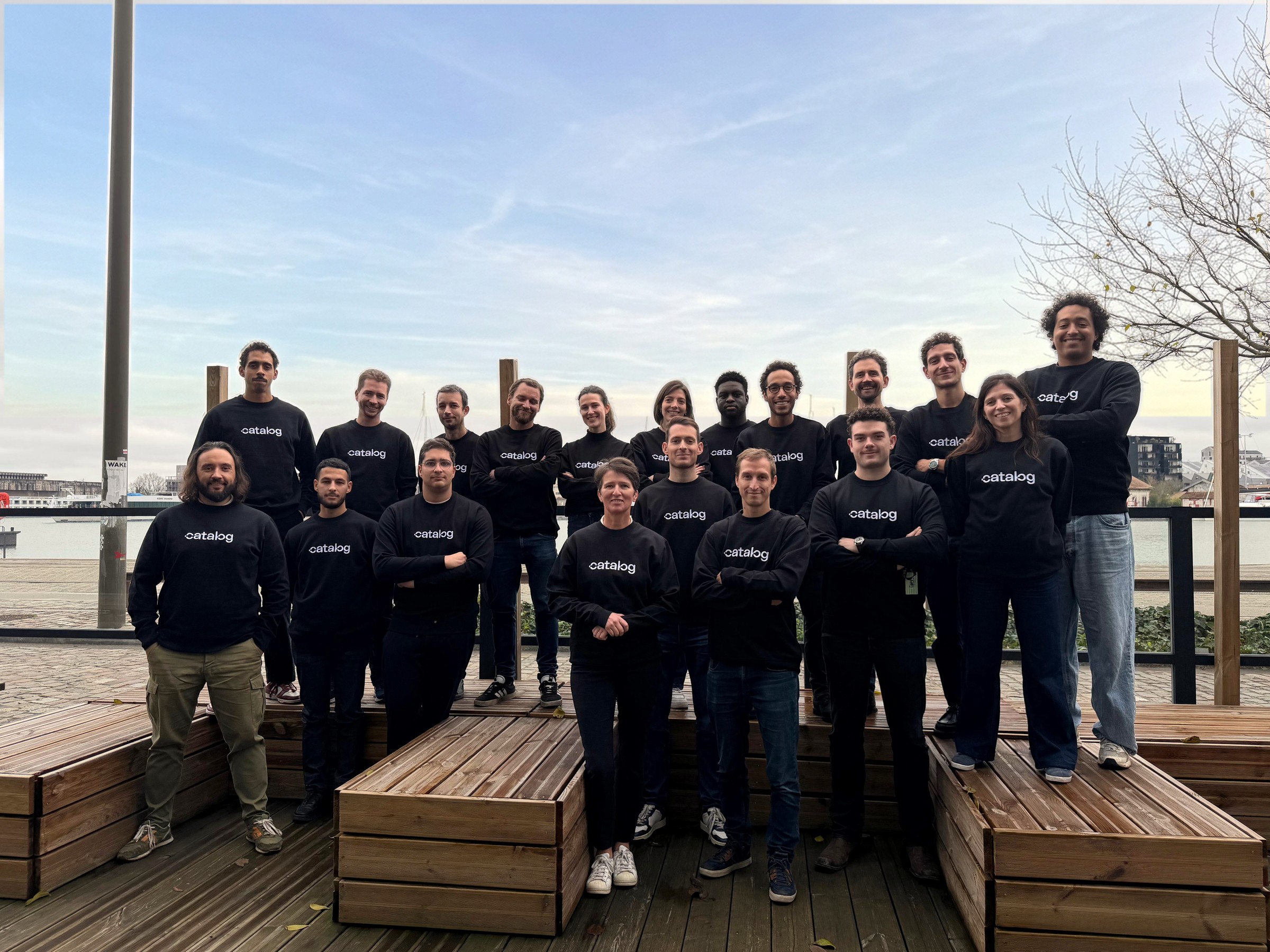 The Catalog team: twenty people in black Catalog sweatshirts on a dock on the Bordeaux waterfront, under a clearing morning sky.